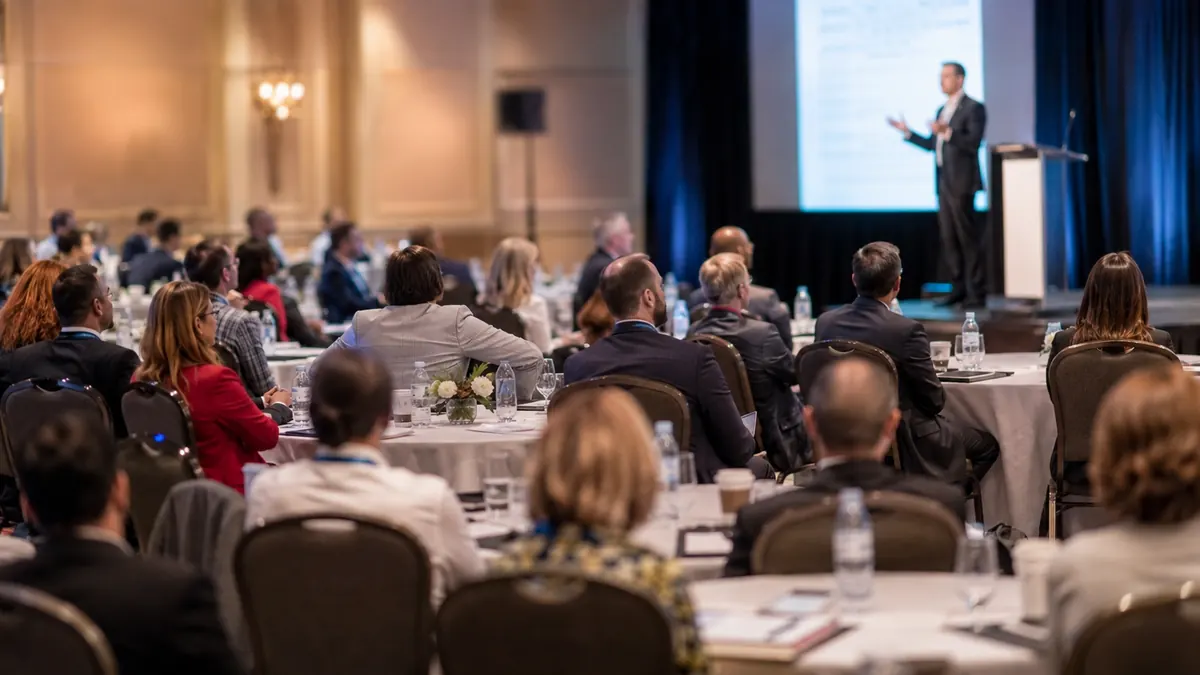 A partner summit: a speaker presenting from a podium to an audience seated at round tables in a ballroom.