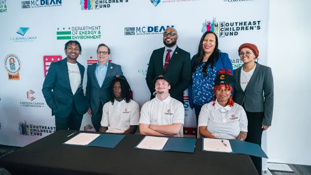 Three Youth Apprenticeship students seated at a signing table with their letters of intent, with programme staff and employer representatives standing behind them at National Signing Day.